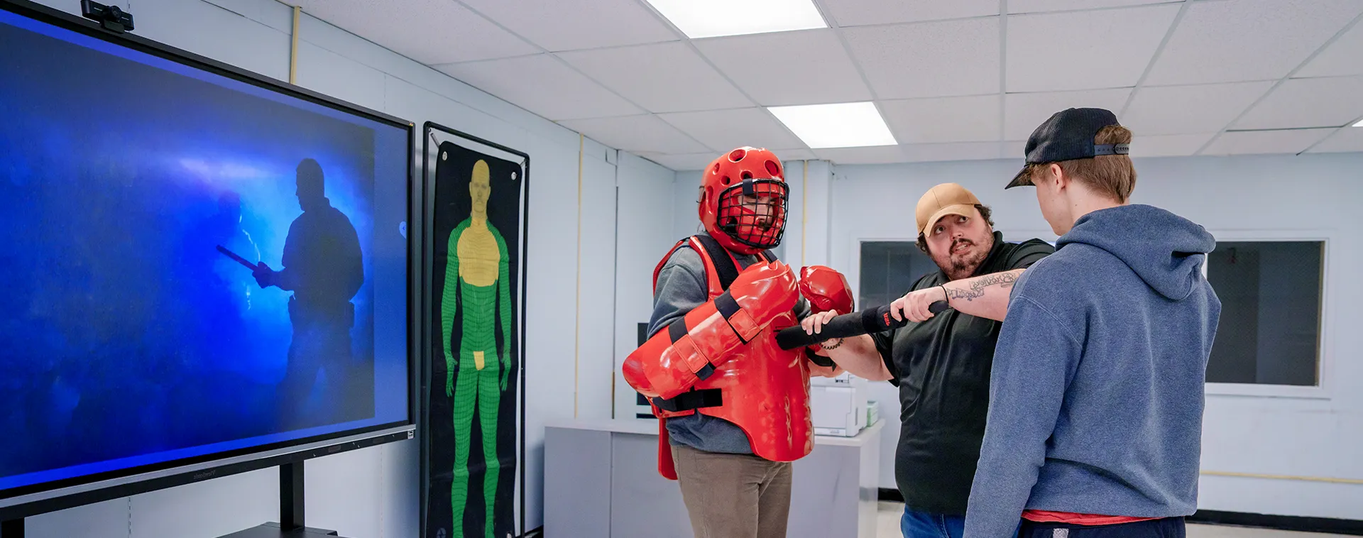Instructor guiding two students in red protective gear during defensive training activity in classroom with wall posters and display screen