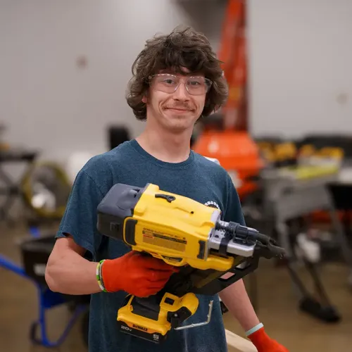 Person wearing safety glasses holding a nail gun in a workshop with tools and equipment in background