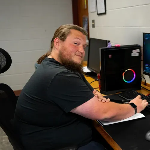 Person seated at desk turning toward camera beside computer with RGB-lit desktop tower and monitor