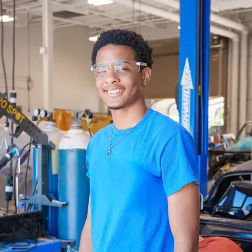 Technician wearing safety glasses standing in auto shop beside gas tanks, tools, and vehicle equipment.