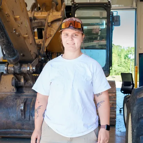 Person standing in front of heavy equipment wearing safety glasses and cap inside a workshop with open doorway in background