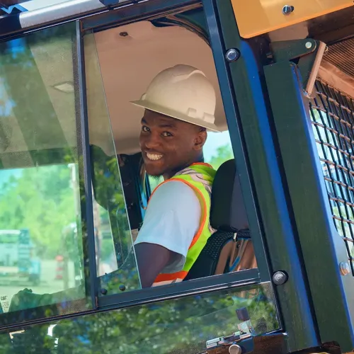 Person wearing hard hat and safety vest seated in equipment cab, smiling toward camera through window
