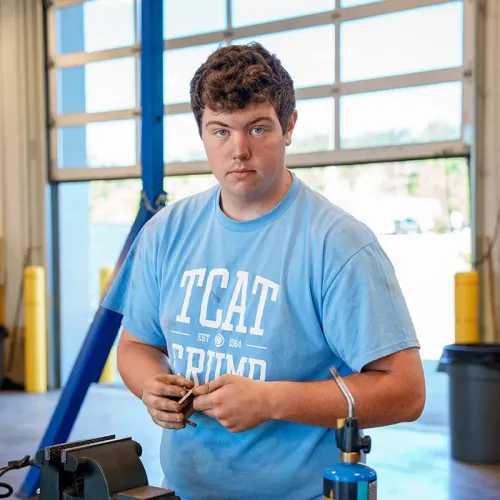 Person in TCAT Crump shirt stands in workshop holding small part near vise and blue torch tank.