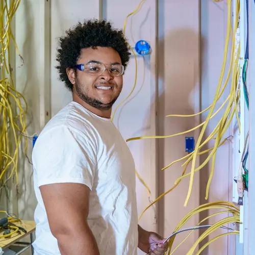 Person wearing safety glasses installs yellow electrical wires into wall studs during wiring practice.