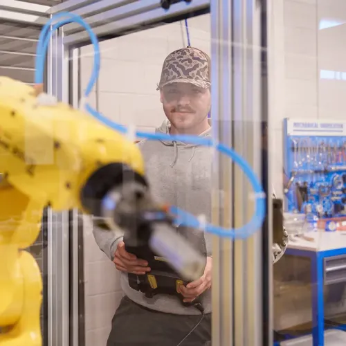 Person stands behind clear enclosure operating yellow robotic arm holding control device in workshop setting.