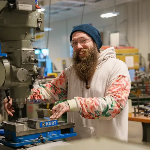 Person in safety glasses operates drill press machine with vise holding metal piece in workshop.