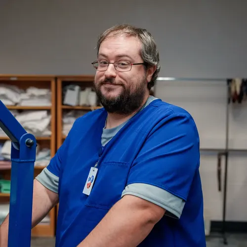 Person in blue scrubs stands beside patient lift in clinical lab with shelves and equipment behind.