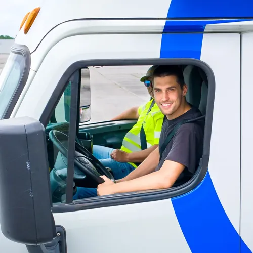 Driver and passenger sit inside semi truck cab; person at wheel looks toward camera through open window.