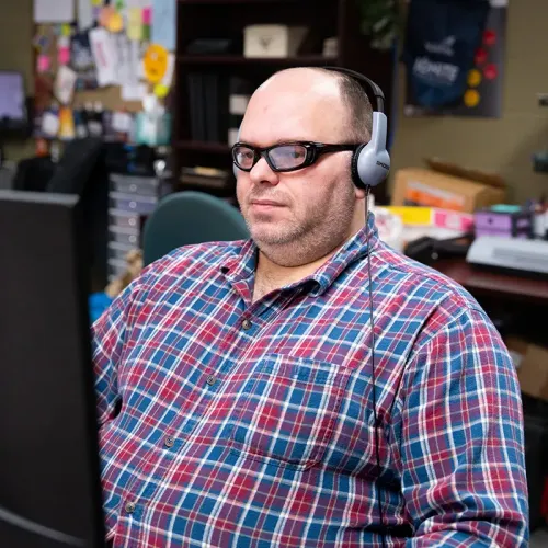 Person wearing headset and glasses working at computer in office with bulletin board and shelves behind.