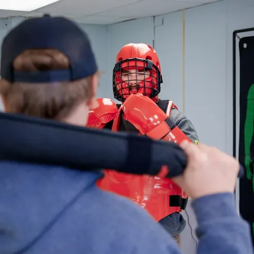 Student holding padded training baton facing person in red protective gear in classroom with white walls and training poster