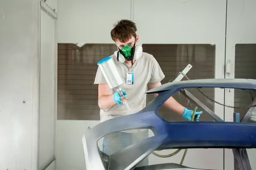 Technician in spray booth wearing respirator and gloves, using paint sprayer on blue car bumper panel.