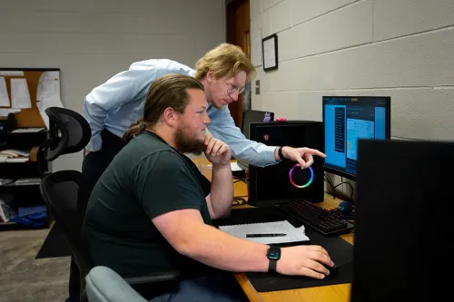 Two people at computer workstation, one pointing at screen while the other uses mouse and keyboard with notebook nearby
