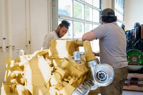 Two people inspecting a large yellow engine with turbocharger in a workshop with tools and machinery around