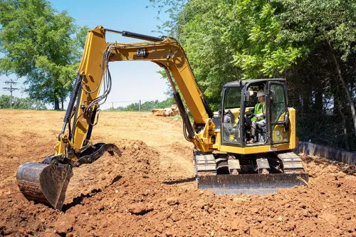 Excavator digging soil on construction site with operator inside cab near tree line