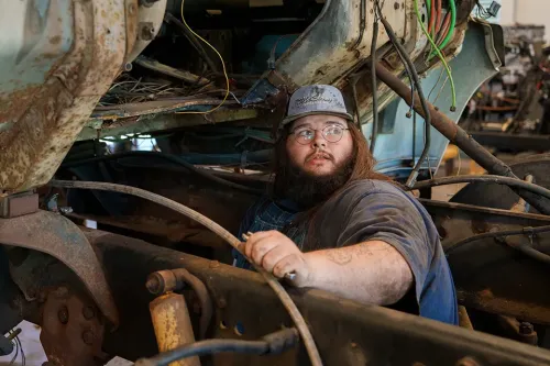 Person in cap works inside open truck engine bay surrounded by wires and metal frame components.