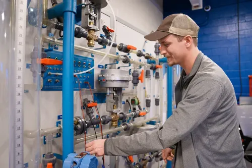 Person adjusts valves and gauges on fluid training board with pipes, meters, and blue wall behind.