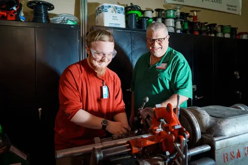 Two people use a pipe threading machine in workshop with spools of wire and cabinets in background.