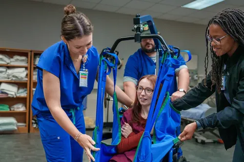 Group uses lift sling to transfer seated person in training lab with shelves of folded linens in background.