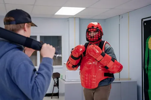 Person wearing red protective training gear and helmet faces another person holding a padded training baton in a classroom.